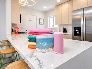 A modern kitchen with a marble countertop, a stack of colorful towels, a guest book, and a pink speaker. - Gorgeous Mountain Sunset Casita with AC Heated Pool Hot Tub Fire pit EV Charger (Palm Springs)
