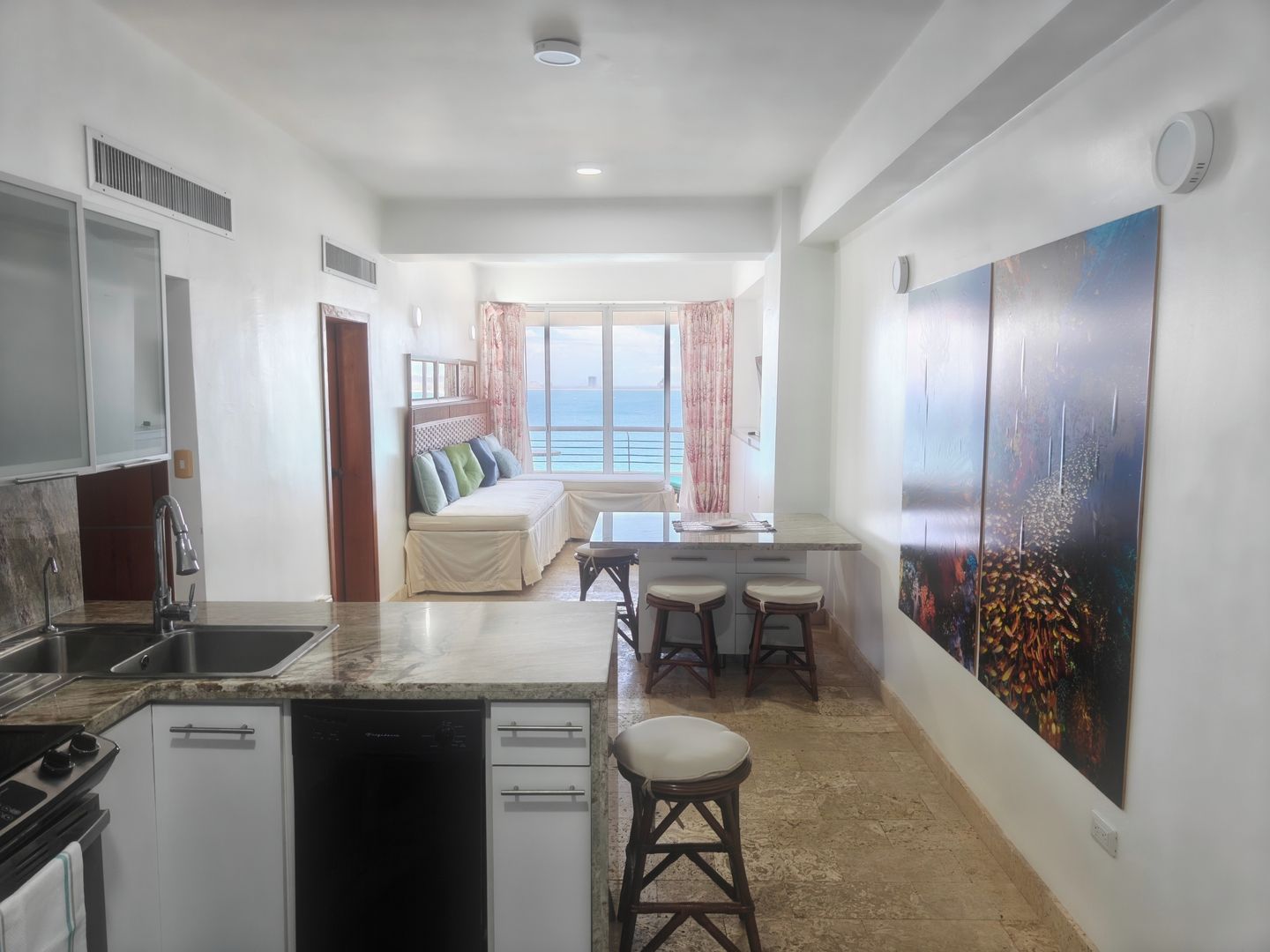 A modern kitchen with a dishwasher, a dining area with a table and two stools, a sofa with a view of the sea, and a large painting on the wall.