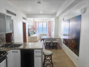 A modern kitchen with a dishwasher, a dining area with a table and two stools, a sofa with a view of the sea, and a large painting on the wall.