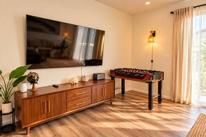A living room with a wooden cabinet, a television mounted on the wall, a foosball table, and a lamp. - Family Friendly Home w/ Parking & Full Kitchen (Long Beach)
