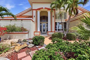 A two-story house with a red and white exterior, a covered front porch, and a landscaped garden with various plants and a rock feature. - Cozy Family House in Punta Gorda, 5919 (Charlotte County)
