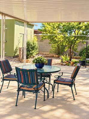 Een terras buiten met een ronde glazen tafel en zes stoelen met gestreepte kussens.