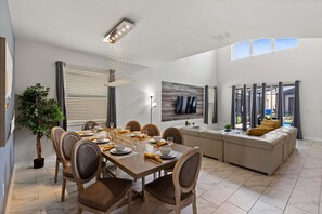 A modern dining area with a wooden table and chairs, a large window with blinds, and a view of the outdoors. - Elegant 5 Bedroom House in Kissimmee, 6701 (Orlando)