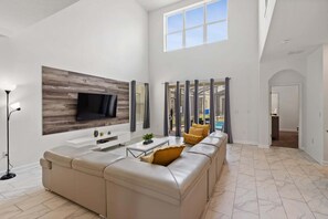 A spacious living room with a large sectional sofa, a flat-screen TV mounted on a wooden panel wall, a coffee table, and a floor lamp. - Elegant 5 Bedroom House in Kissimmee, 6701 (Orlando)