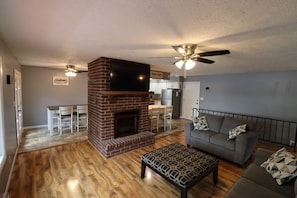 A living room with a brick fireplace, a grey sofa, a patterned ottoman, and a ceiling fan.