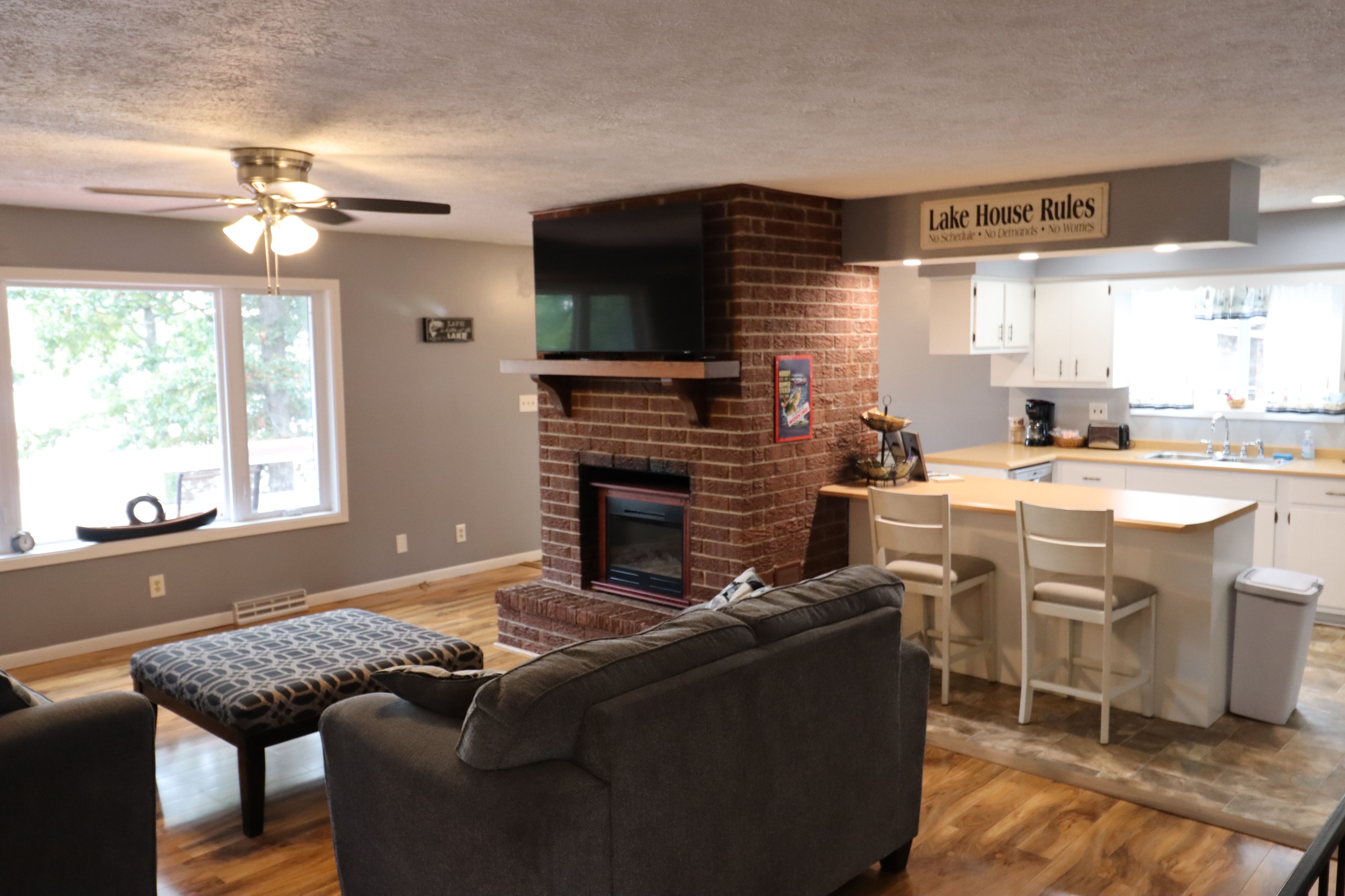 Un salon moderne avec une cheminée, un canapé, un coin repas avec des tabourets de bar et une cuisine sur laquelle est affiché un panneau indiquant : « Règles de la maison au bord du lac ».