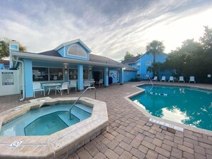 Un área de piscina con jacuzzi y sillas de descanso, un espacio de estar cubierto, y un edificio con fachada azul.