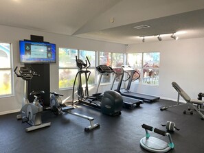 A gym with various exercise equipment, a TV displaying sports scores, and large windows with a view of the outdoors. - Cozy 3 Bedroom Condo for Sale in Kissimmee, 6699 (Orlando)