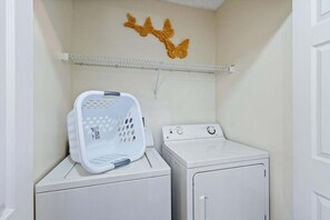 A laundry area with a washing machine and dryer, a white laundry basket, and a shelf with decorative butterfly wall art. - Cozy 3 Bedroom Condo for Sale in Kissimmee, 6699 (Orlando)