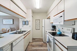 Cocina compacta con muebles blancos, lavavajillas, estufa y microondas. Hay citas enmarcadas en la pared y un adorno sobre el lavamanos.