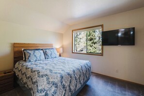 A bedroom with a bed, a TV mounted on the wall, and a window with a view of trees. - Sunriver Fun - Fireplaces, Hot tub & SHARC (Sunriver)