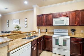 A kitchen with granite countertops, wooden cabinets, a white microwave, a stove, and a dishwasher. - Serene Retreat: 2 Bed Condo in Davenport, 6707 (Orlando)