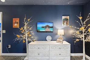 Een kamer met een donkerblauwe wand, een televisie waarop een strandscène te zien is, een witte ladekast en decoratieve elementen, waaronder een bordje met de tekst 'LIFE IS BETTER at the Campsite' en een 'CANONES' bord.