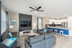 A modern living room with a sofa, coffee table, and TV, a kitchen with white cabinets, and a ceiling fan. - Stunning 10 Bedroom Home in Davenport, 6705 (Orlando)