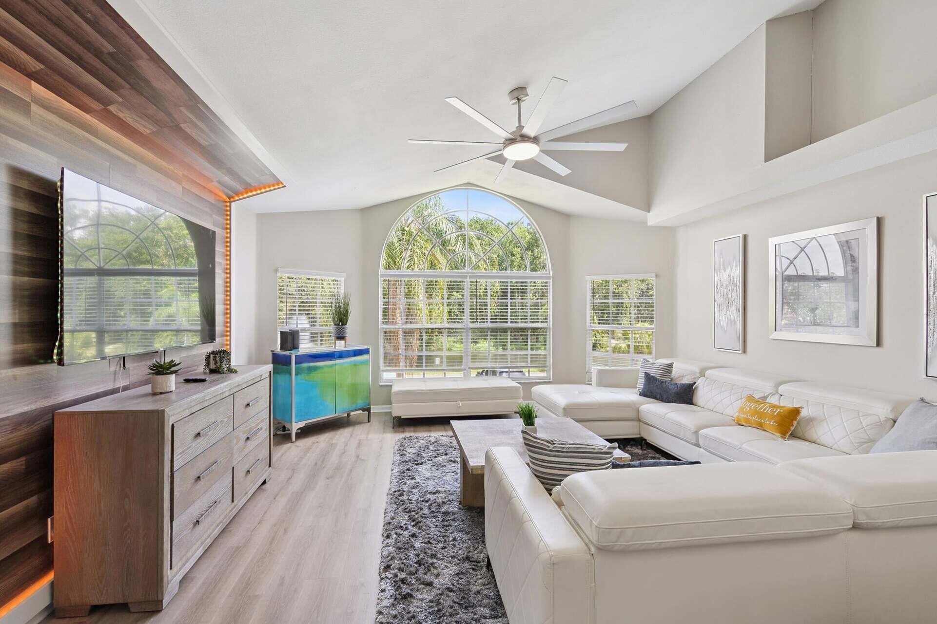 A spacious living room with a high ceiling fan, large windows, a white sofa, and a wooden cabinet.