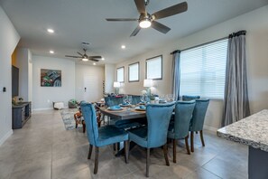A dining area with a table set for six, surrounded by six blue chairs, a ceiling fan, and a window with blinds. - Inviting Family Home in Davenport Area, 6695 (Orlando)