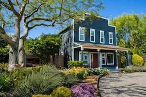 A two-story house with a porch, surrounded by a garden with various flowers and a large tree.