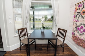 A dining area with a dark wooden table and two chairs, a potted plant, and a view through large windows with blinds.