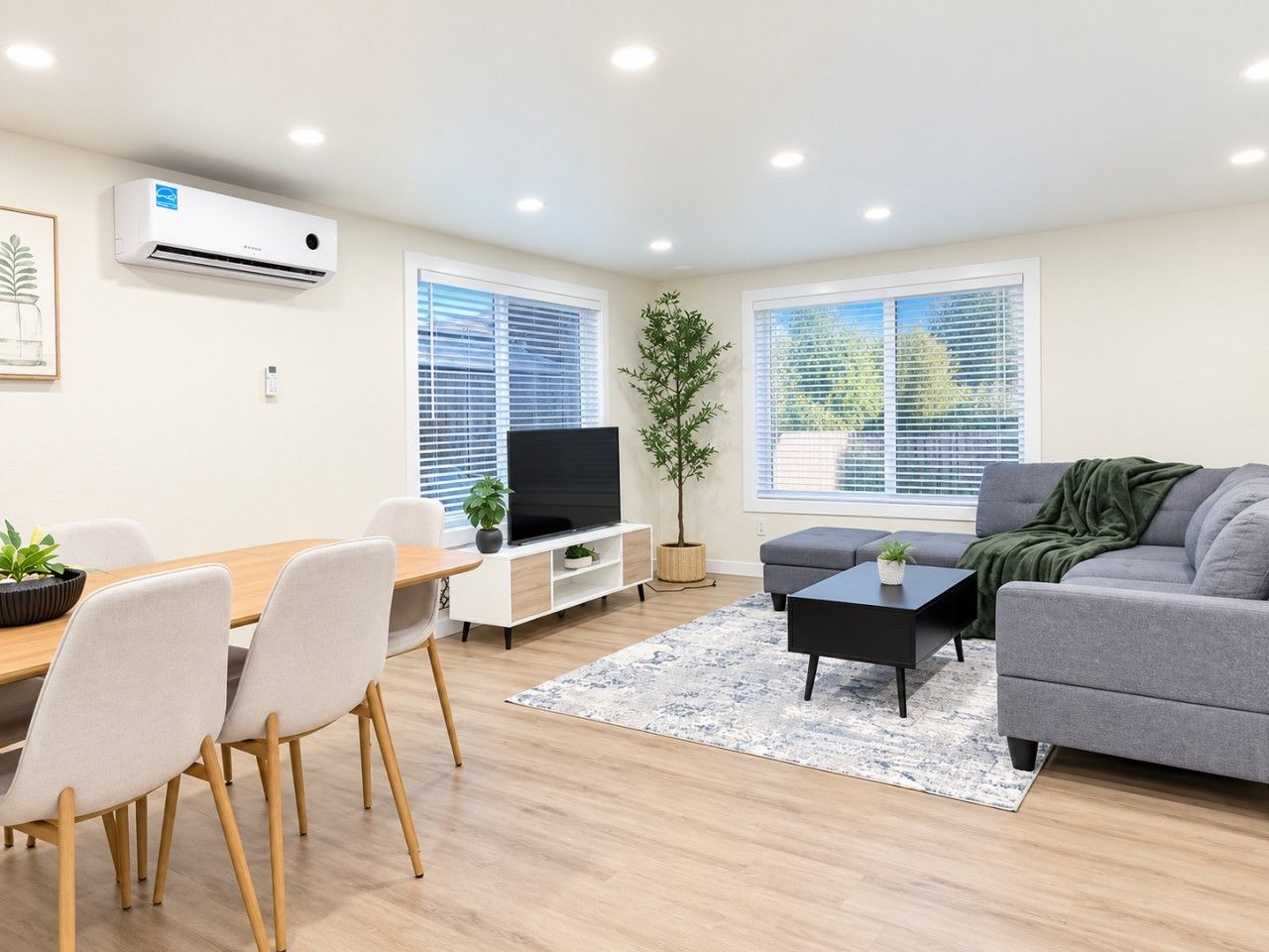 Een moderne woonkamer met een grijze bank, een zwarte salontafel, een televisie en een eethoek met een houten tafel en stoelen.