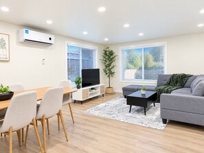 A modern living room with a grey sofa, a black coffee table, a TV, and a dining area with a wooden table and chairs.