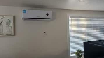 A room with a mounted air conditioner, a window with blinds, a flat-screen TV, and a small plant on the windowsill.