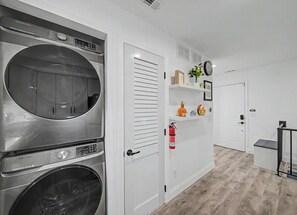 A laundry room with two front-loading washing machines, a white door with a handle, a fire extinguisher mounted on the wall, and a small shelf with decorative items. - Mountain Creek Ski Condo | 3BR, Arcade, Mtn Views (Vernon Township)