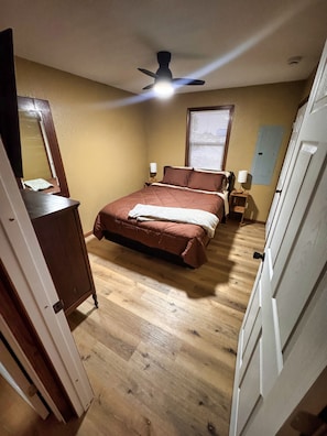 A bedroom with a bed, a ceiling fan, a window with blinds, and a wooden floor.