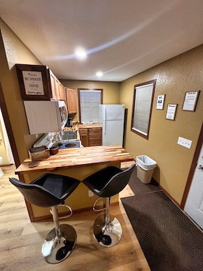A compact kitchen with wooden cabinets, a white refrigerator, a wooden countertop, two bar stools, and a trash can.