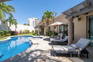 A swimming pool area with lounge chairs and umbrellas, surrounded by palm trees and a building in the background. - A 100% Private Home for Family and Friends (San José del Cabo)