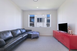 A room with a black leather sofa, a red cabinet with a television, and a window with blinds. - Wooloowin Escape Modern Comfort and Sunlit Courtyard (Brisbane)