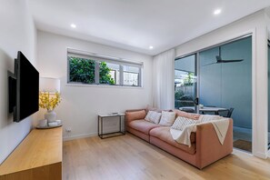 Ein modernes Wohnzimmer mit einer Couch, einem Flachbildfernseher, einem kleinen Tisch mit einer Lampe und einem Fenster mit Blick auf Grünflächen.