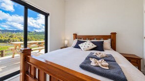 A bedroom with a wooden bed, pillows, a bedside lamp, and a view of a park through a large window. - Road to Paradise | Mountain Views & Tranquility (Kopu)