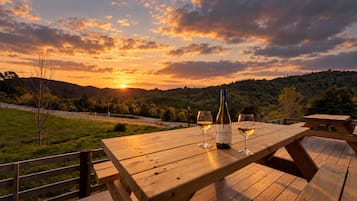 Una mesa de picnic de madera con una botella de vino y dos copas, frente a una vista al atardecer con una carretera y colinas de fondo.