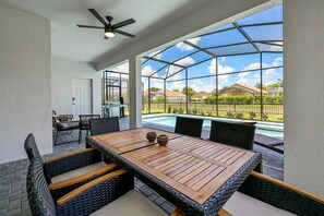 Une terrasse extérieure avec une table en bois et des chaises en osier, un ventilateur de plafond, et une vue sur une piscine et des maisons en arrière-plan.