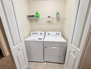 A laundry room with two white front-loading washers and dryers, a metal shelf above holding laundry supplies, and beige carpeted floor. - 5BR Disney-Themed Getaway with Pool (Davenport)