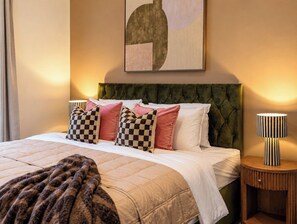 Quarto de hotel com cama, cabeceira estofada em verde, almofadas xadrez, manta e luminária em uma mesinha de madeira.