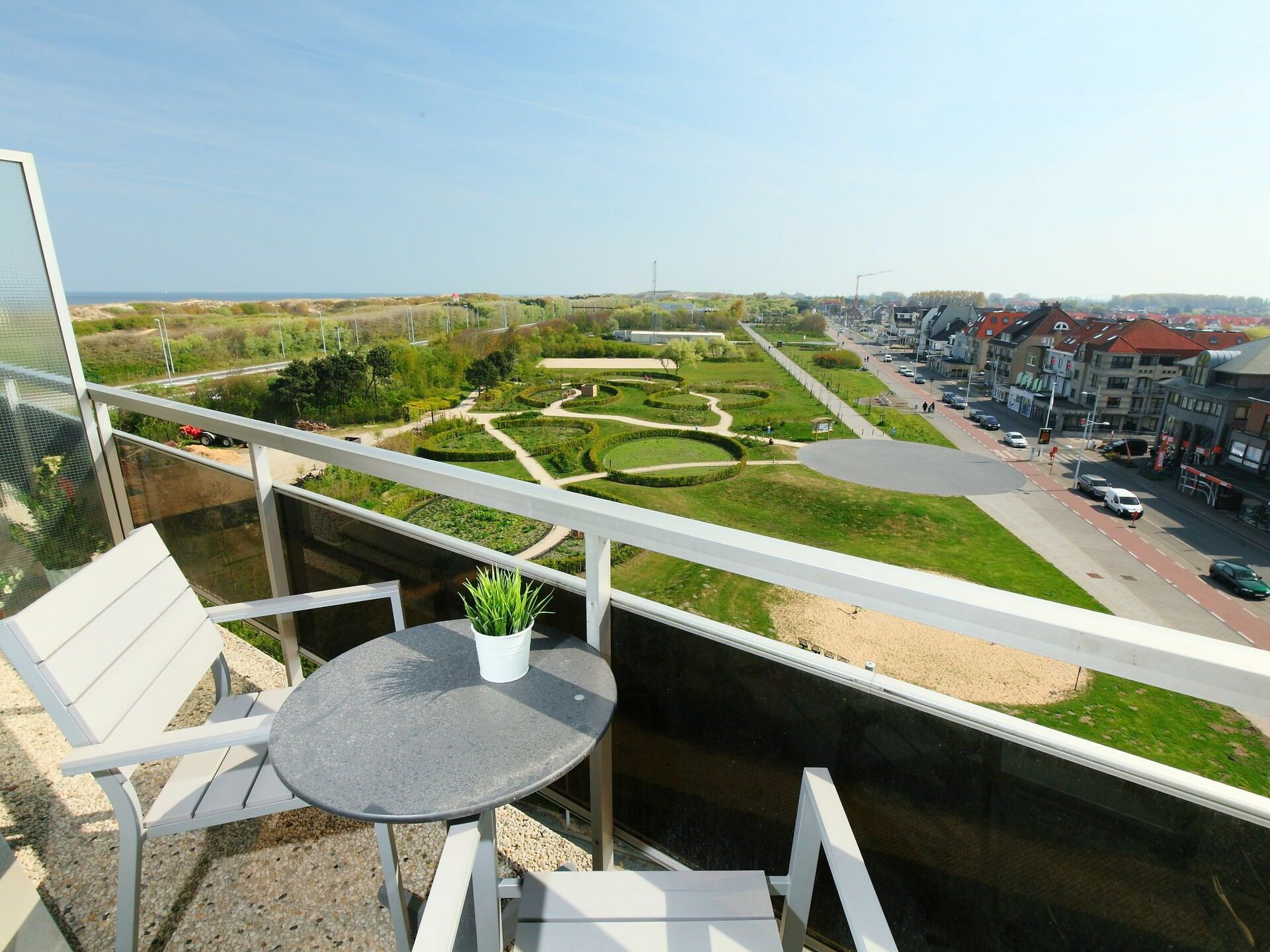 A balcony with a table and chairs, overlooking a landscaped garden and a street with buildings.
