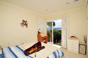 A bedroom with a bed, a violin, a small wooden cabinet, and a view of greenery through a sliding glass door. - Moana View - Elevated Beachside Home with Panoramic Ocean Views (Nelson)