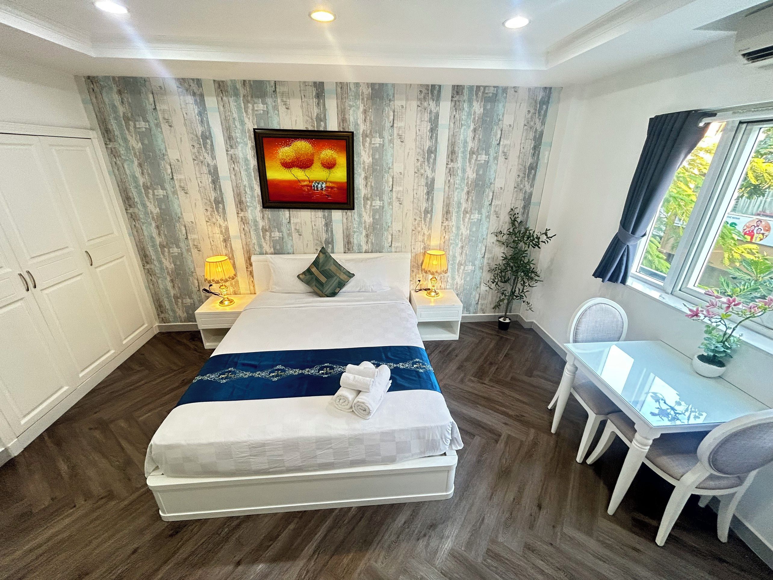 A hotel room with a bed, bedside lamps, a desk with chairs, a window with curtains, and a wall-mounted TV.
