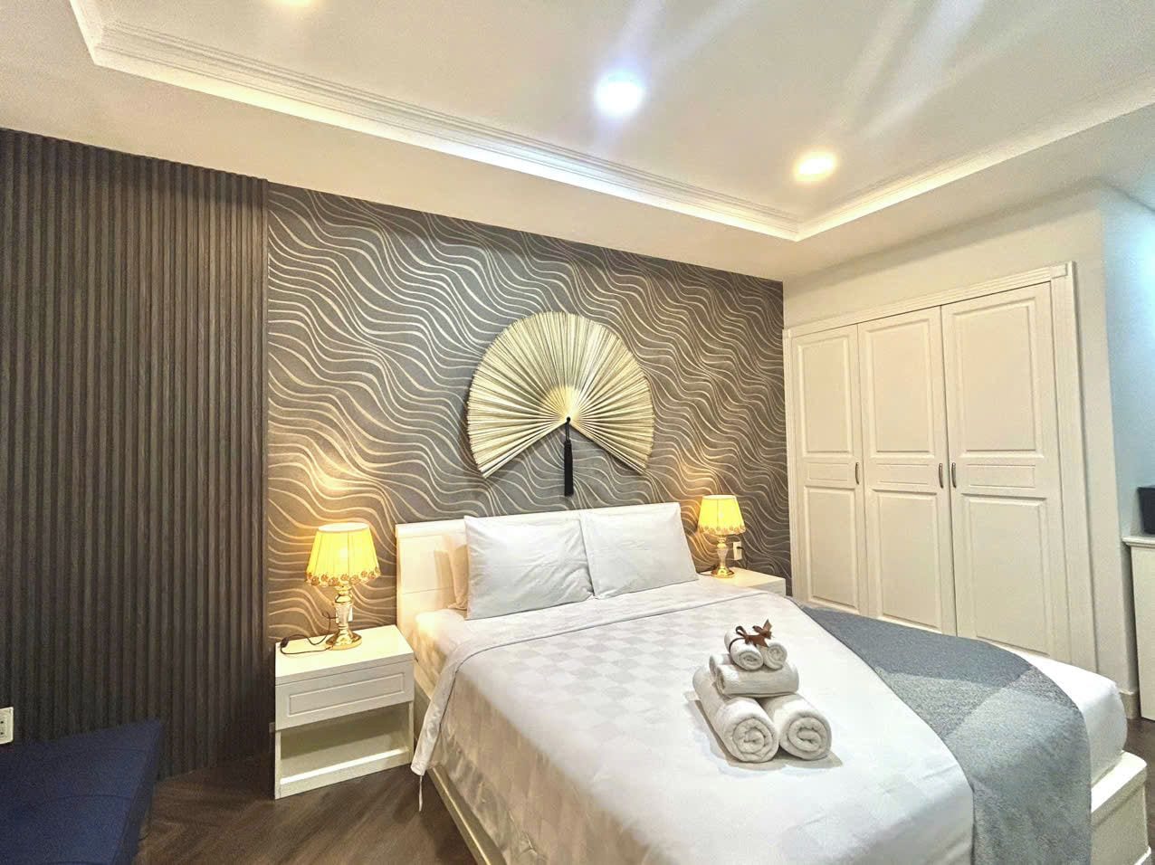 A hotel room with a bed, two bedside lamps, a wall-mounted fan, and a wardrobe.