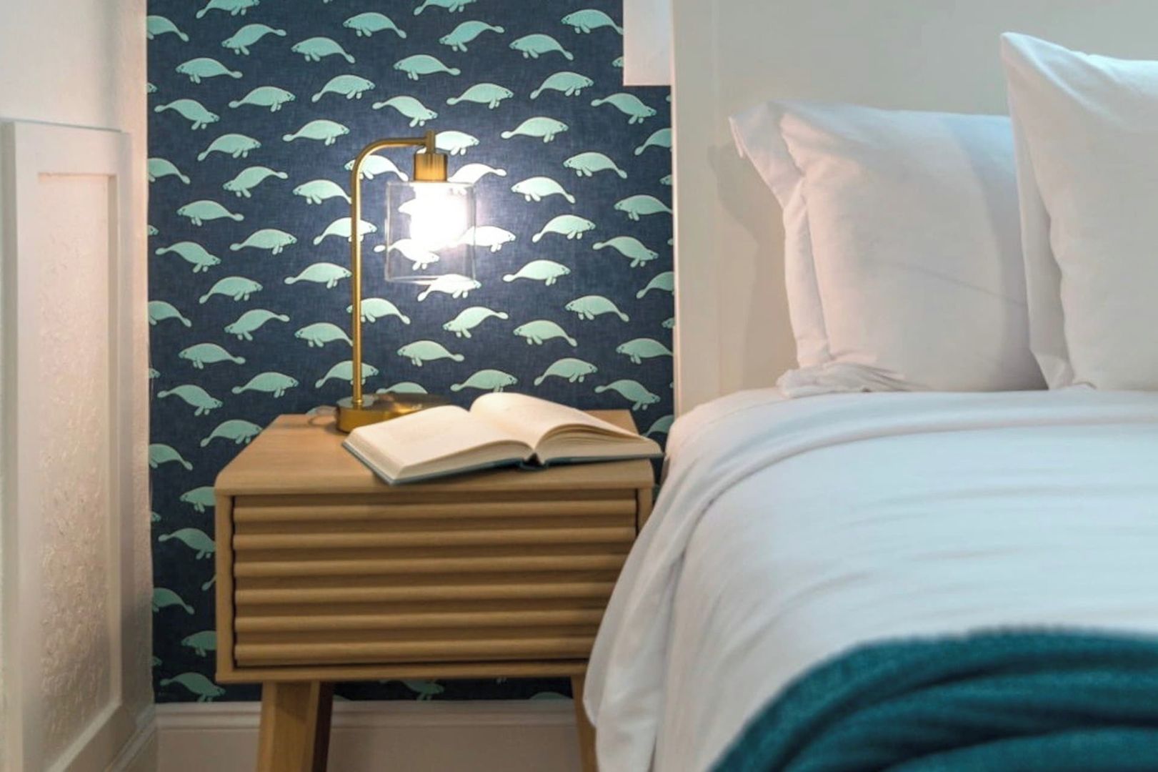 A bedroom with a bed, pillows, a bedside table with an open book, a lamp, and a wall with a pattern of sea creatures.