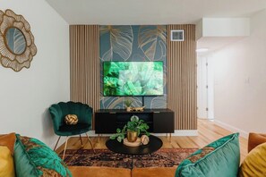 Un salon doté d’un téléviseur à écran plat fixé au mur, lequel est orné d’un motif de feuilles, d’un meuble de rangement sombre, d’une table basse ronde agrémentée d’une plante et de deux fauteuils verts.