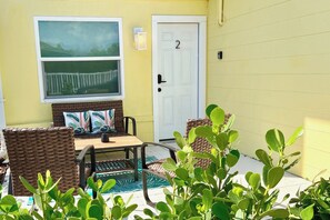 Un bâtiment jaune avec une porte portant le numéro « 2 », une fenêtre donnant sur une clôture, et du mobilier d’extérieur comprenant une table et des chaises.