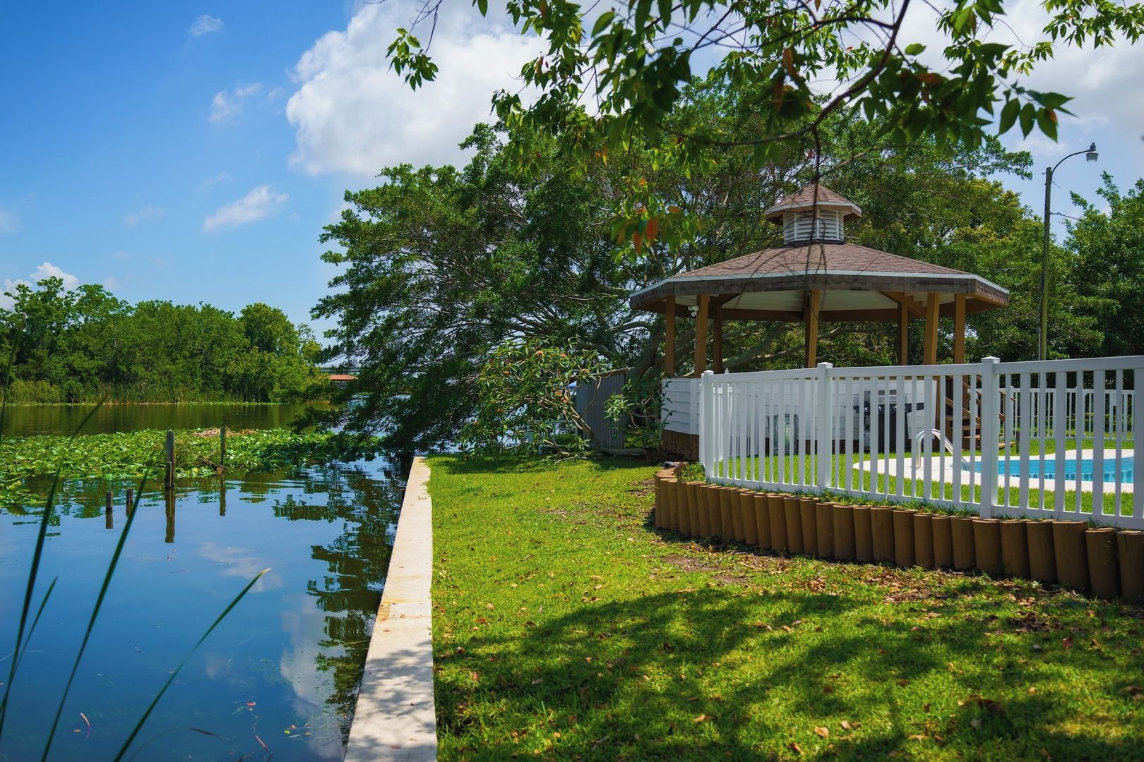 A gazebo with a white fence, a swimming pool, and a body of water with lily pads.