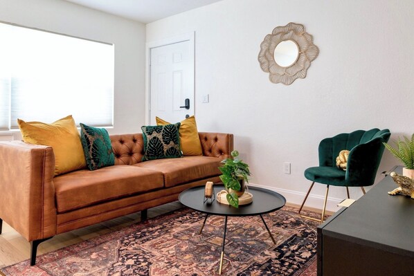 Una sala de estar con un sofá marrón, una mesa redonda de centro, un sillón verde, un tapete estampado y un espejo decorativo.
