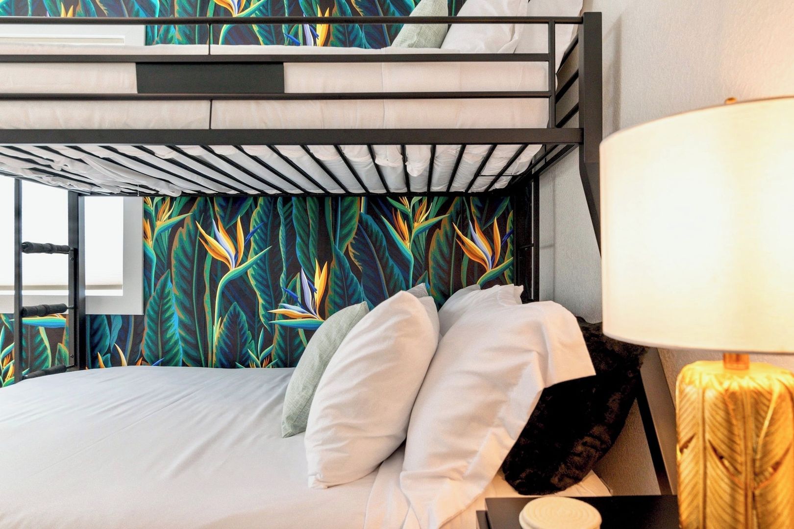 A bunk bed with a tropical leaf pattern wall, white bedding, and a bedside lamp.