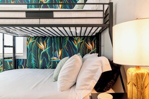 Un lit superposé avec un mur décoré d’un motif de feuilles tropicales, du linge de lit blanc et une lampe de chevet.
