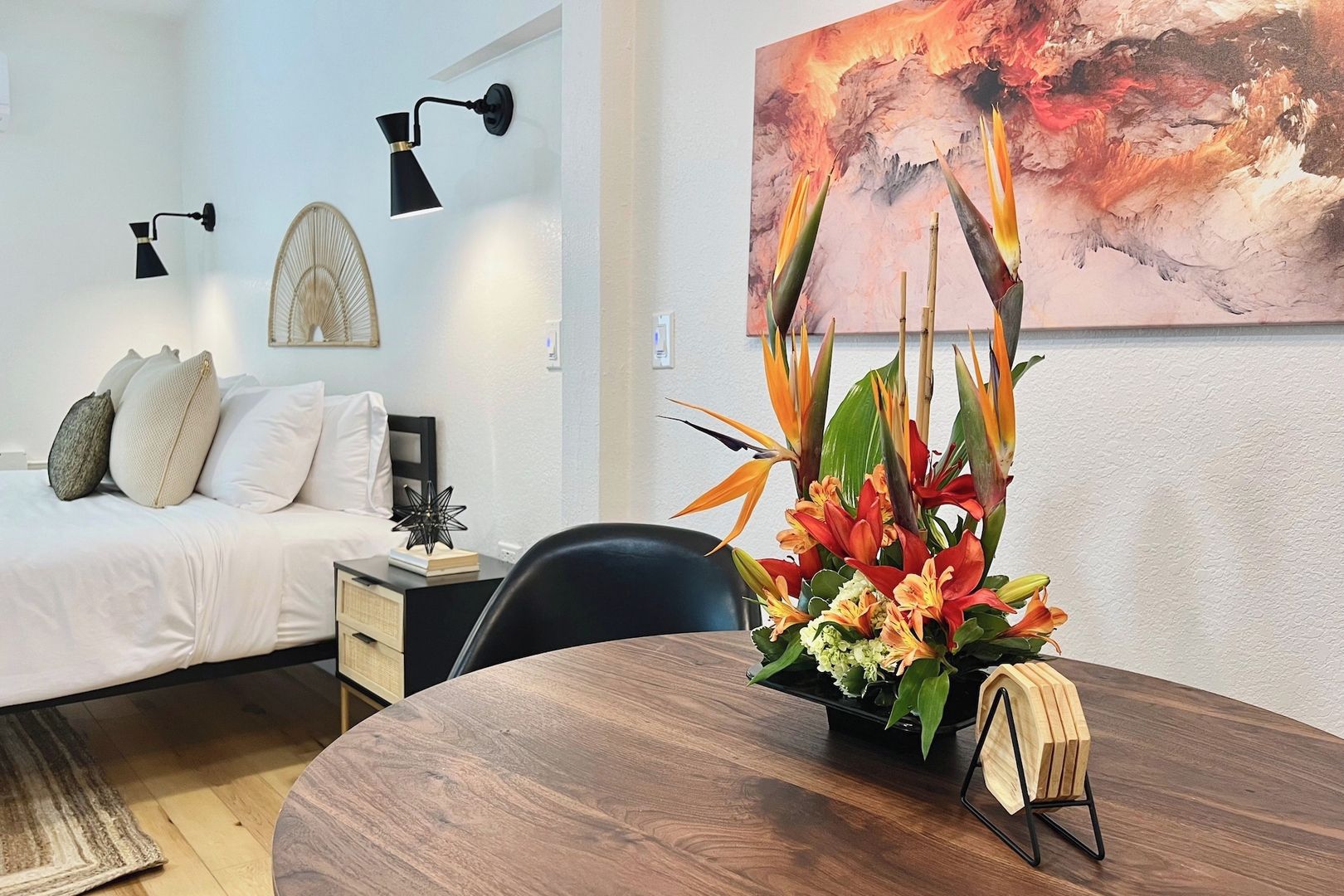 A hotel room with a bed, bedside lamps, a desk with a chair, a dining table with a flower arrangement, and a painting on the wall.