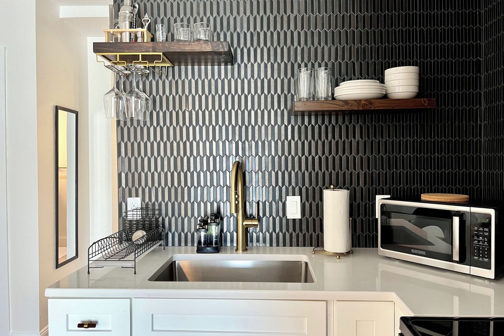 A modern kitchen with a geometric patterned backsplash, stainless steel appliances, and a wooden shelf with glassware.