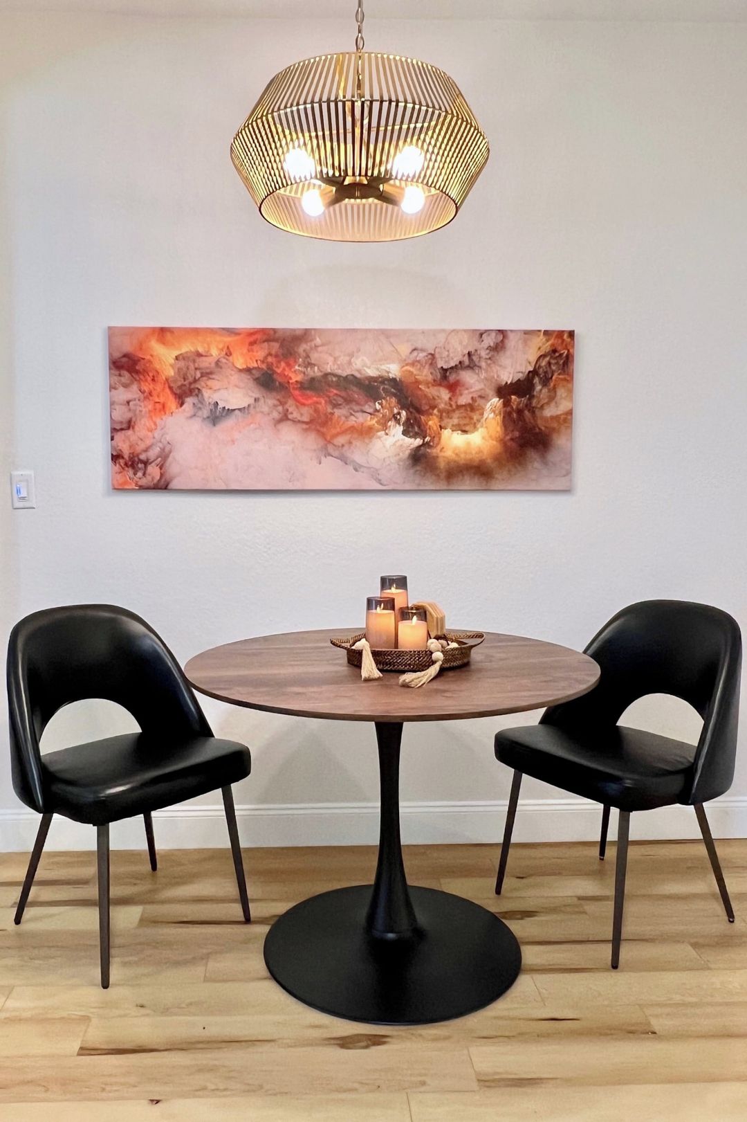 Un coin repas comprenant une table ronde, deux chaises, un luminaire suspendu et un grand tableau abstrait accroché au mur.
